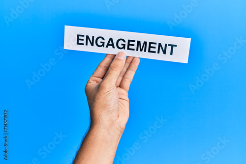 Hand of caucasian man holding paper with engagement word over isolated blue background