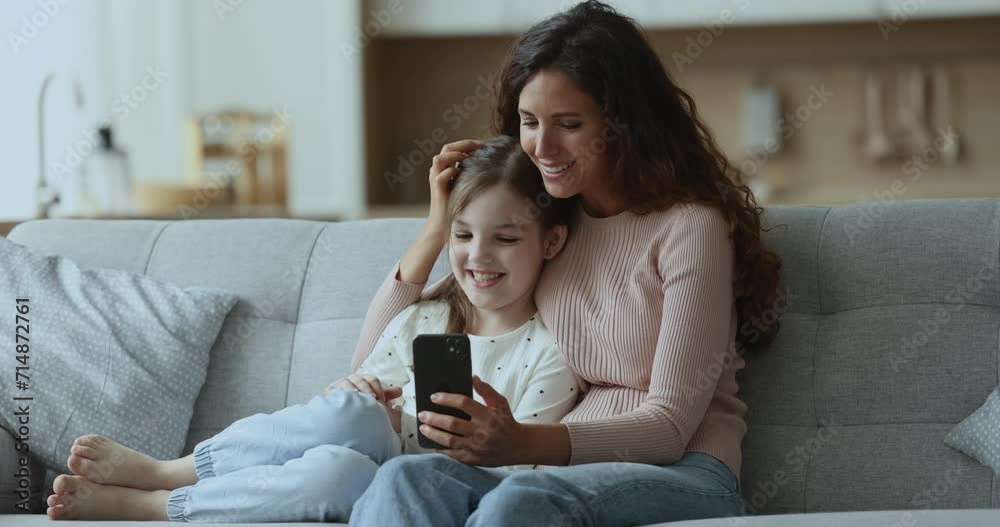 Cheerful beautiful Latin mom and cute daughter girl watching online video content on smartphone, hugging on comfortable home couch, using mobile phone for online entertainment Cheerful beautiful Latin mom and cute daughter girl watching online video content on smartphone, hugging on comfortable home couch, using mobile phone for online entertainment