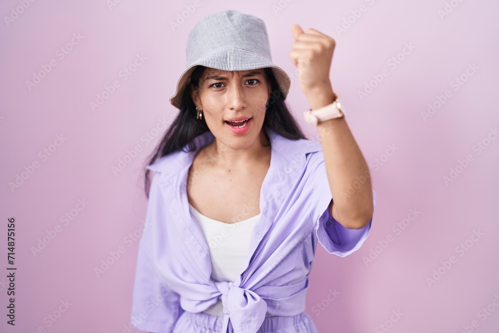 Young hispanic woman standing over pink background wearing hat angry and mad raising fist frustrated and furious while shouting with anger. rage and aggressive concept. Young hispanic woman standing over pink background wearing hat angry and mad raising fist frustrated and furious while shouting with anger. rage and aggressive concept.