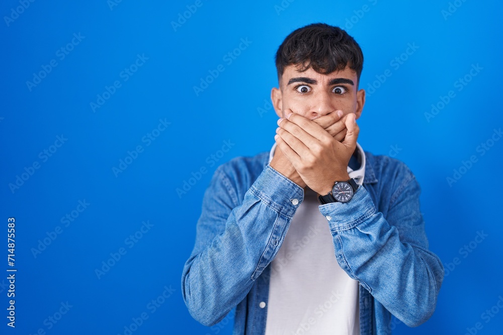Young hispanic man standing over blue background shocked covering mouth with hands for mistake. secret concept.