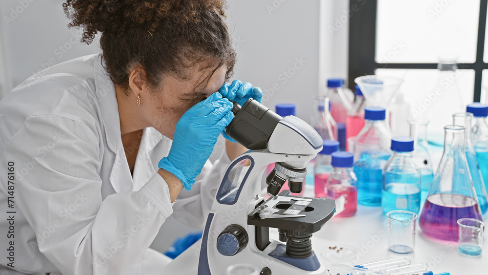 Hispanic woman with curly hair using microscope in laboratory Stock
