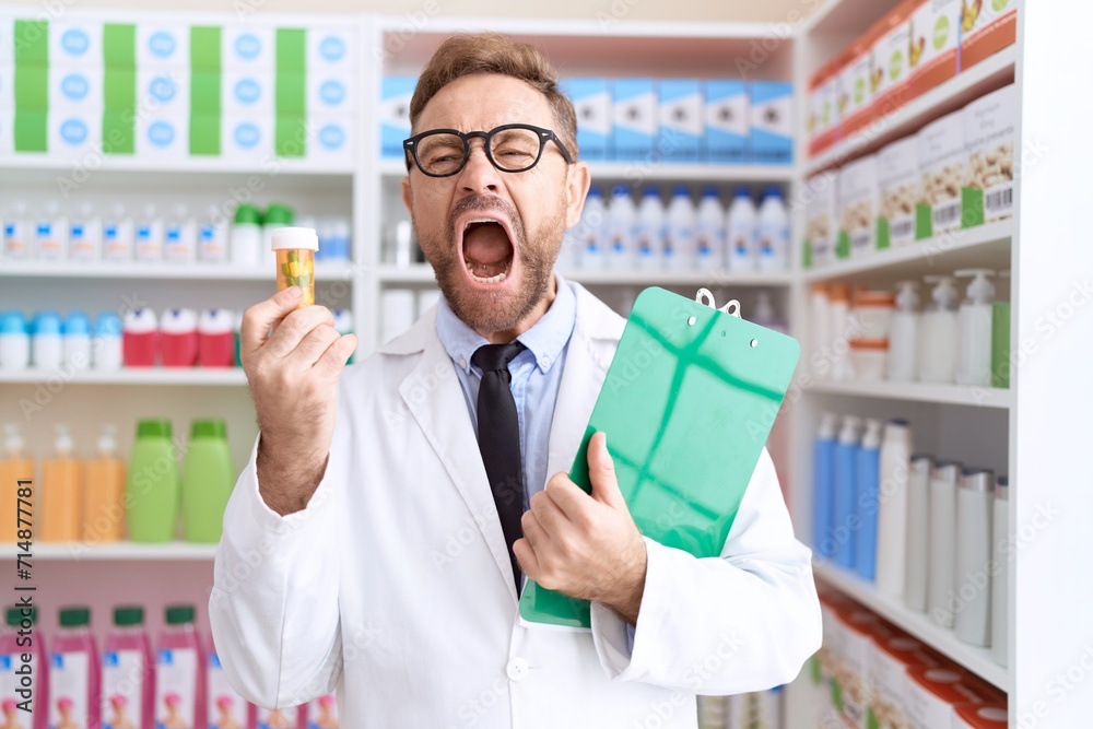 Middle age man with beard working at pharmacy drugstore holding pills angry and mad screaming frustrated and furious, shouting with anger. rage and aggressive concept.