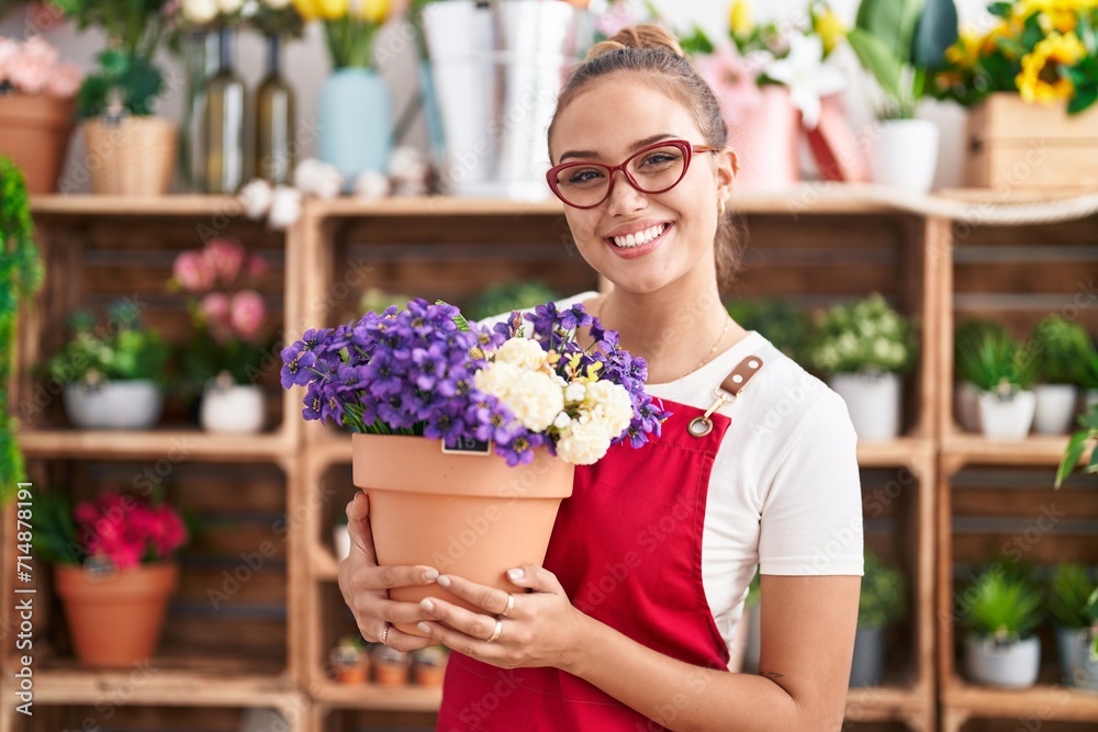 Young hispanic woman working at florist shop holding plant looking positive and happy standing and smiling with a confident smile showing teeth