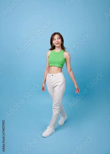 Full body smiling happy young Asian teen woman walking going strolling and looking sideways isolated on blue background studio portrait.