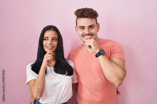 Young hispanic couple standing over pink background with hand on chin thinking about question, pensive expression. smiling and thoughtful face. doubt concept.