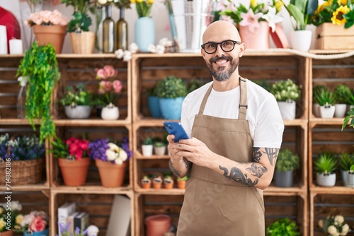 Young bald man florist smiling confident using smartphone at florist