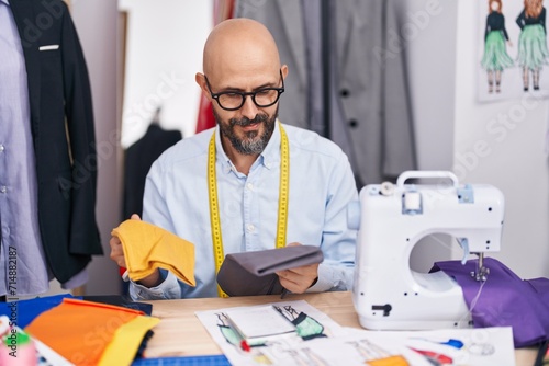 Young bald man tailor smiling confident holding cloths at tailor shop