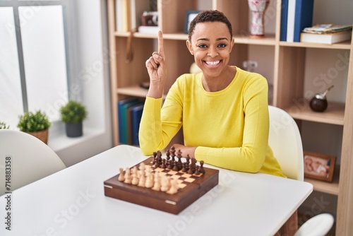 African american woman playing chess sitting on the table smiling with an idea or question pointing finger with happy face, number one