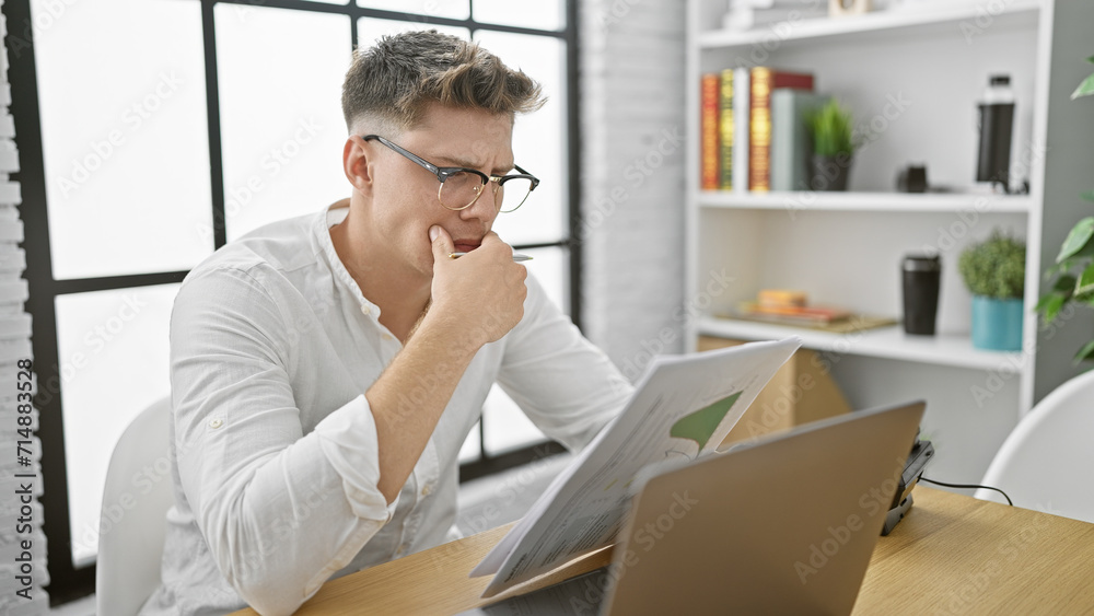 Handsome, bearded, young caucasian man, in glasses, seriously engrossed in reading a business document inside his office, a picture of focused professional in work mode. Handsome, bearded, young caucasian man, in glasses, seriously engrossed in reading a business document inside his office, a picture of focused professional in work mode.