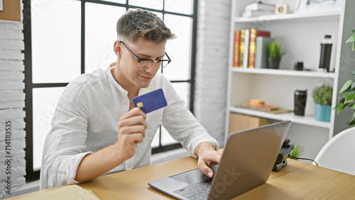 Confident young caucasian man smiles, happily shopping online with credit card at office, working hard as successful manager