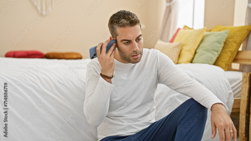 Handsome hispanic man listening to a voice message in a cozy bedroom setting, portraying everyday home life.
