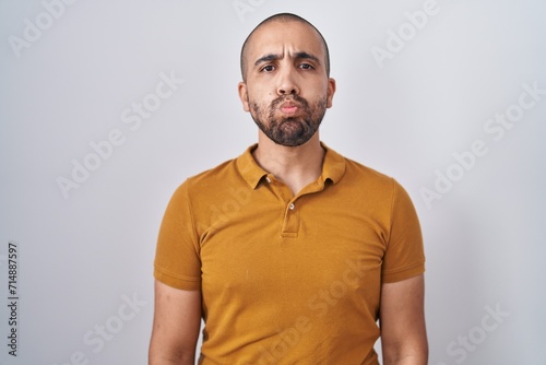 Hispanic man with beard standing over white background puffing cheeks with funny face. mouth inflated with air, crazy expression. photo