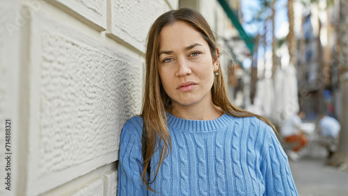 Young woman with long brunette hair and blue eyes wearing a blue sweater posing thoughtfully on a city street.