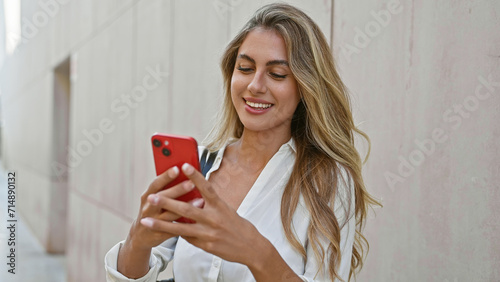 Beautiful young blonde woman tapping into happiness, deeply engrossed in an exciting online conversation on her smartphone texting and smiling outdoors on urban street, radiating cheerful vibes