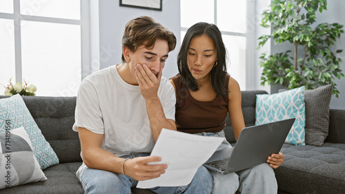 Fotografi Interracial couple analyzes documents in a modern living room with a laptop, expressing concern and teamwork