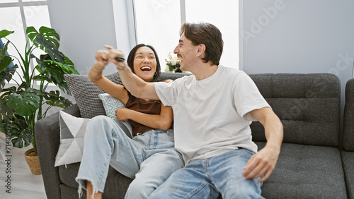 A smiling interracial couple, a woman and a man, laughing and holding a remote control in a cozy living room.