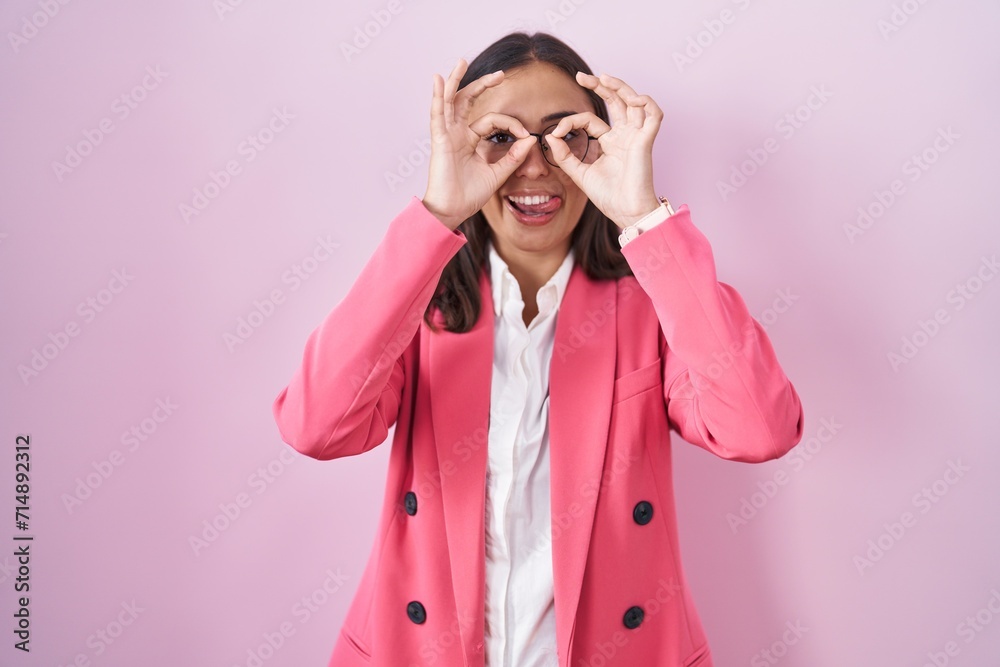Young hispanic woman wearing business clothes and glasses doing ok gesture like binoculars sticking tongue out, eyes looking through fingers. crazy expression. Young hispanic woman wearing business clothes and glasses doing ok gesture like binoculars sticking tongue out, eyes looking through fingers. crazy expression.