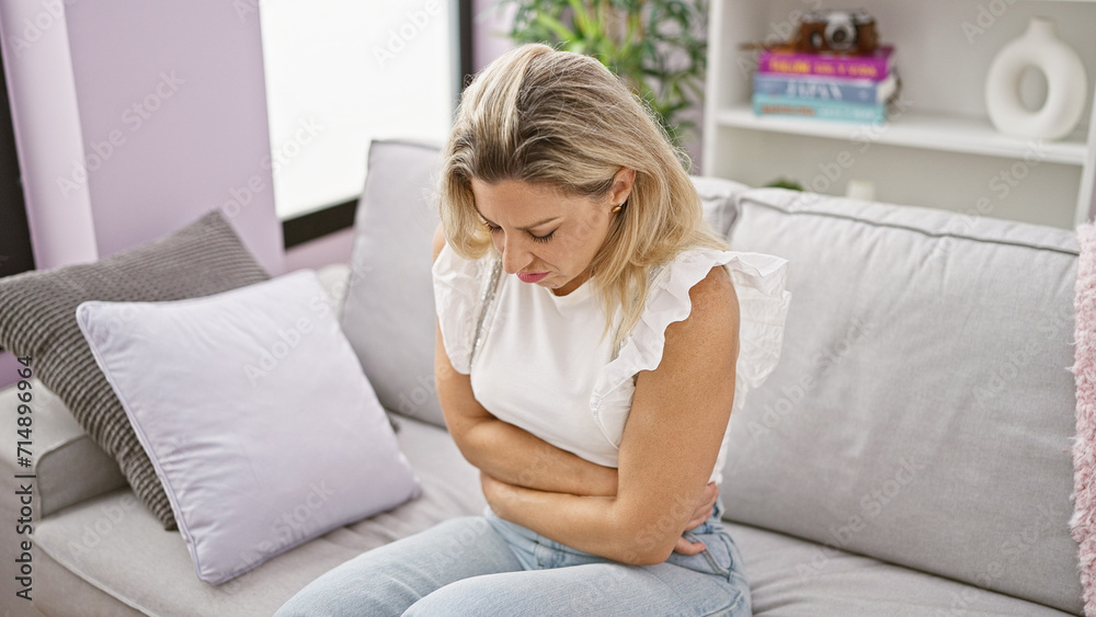 Young blonde woman suffering for stomach ache sitting on sofa at home Young blonde woman suffering for stomach ache sitting on sofa at home