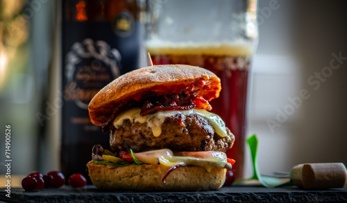 Homemade bacon cheeseburger on a dark tile with berries on the side and cold beer in the background. Food porn and nasty burger, smash burger photo