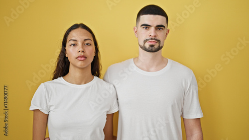 Beautiful couple standing together with serious face over isolated yellow background