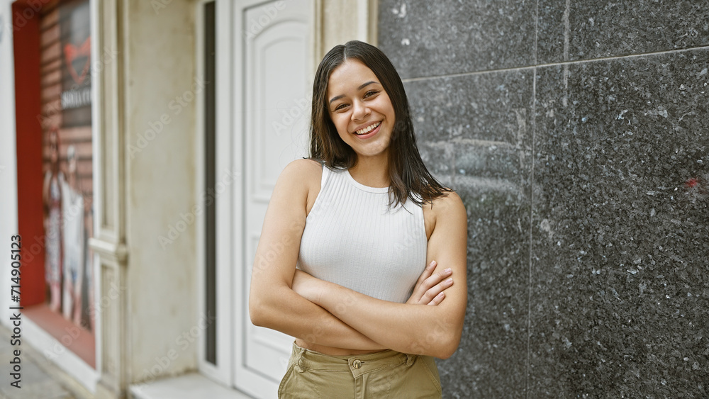 Confident and beautiful - a young hispanic woman radiating positive joy, smiling broadly with arms crossed, stands on a lively city street, embodying an urban, casual chic lifestyle