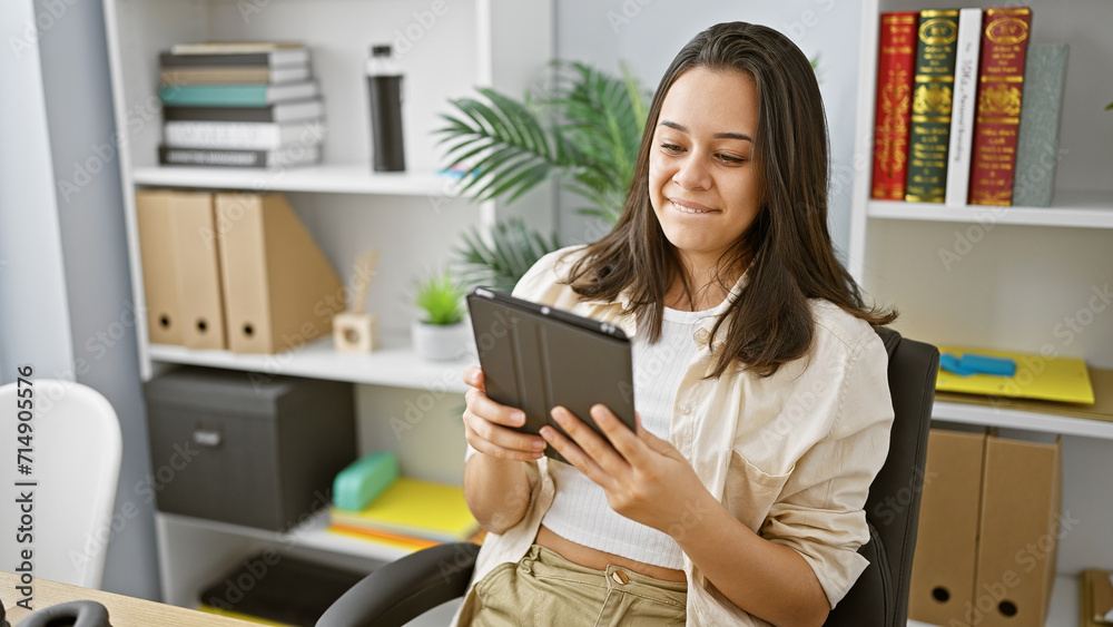 Confident young hispanic woman, a beautiful smiling business worker, enjoying success as she uses touchpad at her elegant office desk indoors Confident young hispanic woman, a beautiful smiling business worker, enjoying success as she uses touchpad at her elegant office desk indoors