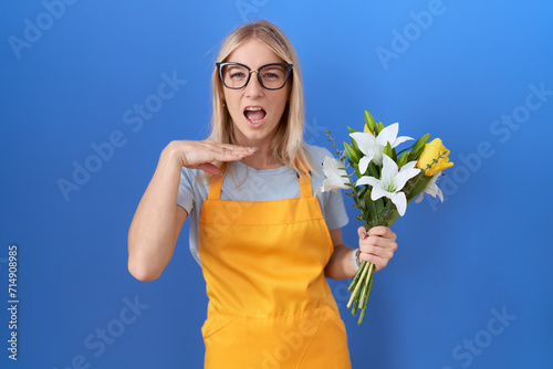 Young caucasian woman wearing florist apron holding flowers cutting throat with hand as knife  threaten aggression with furious violence