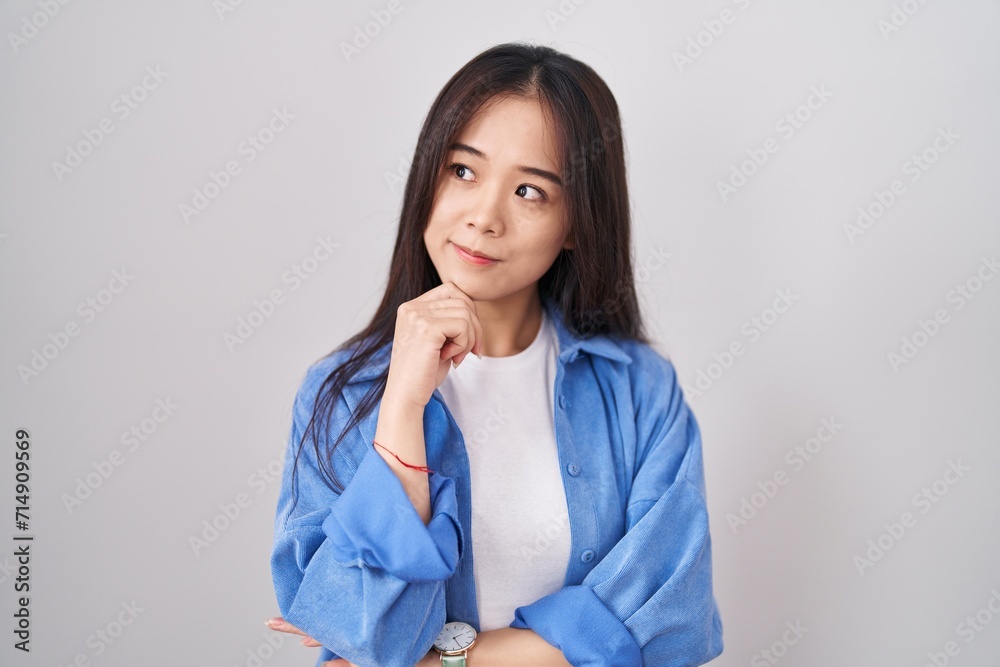 Young chinese woman standing over white background with hand on chin thinking about question, pensive expression. smiling with thoughtful face. doubt concept. Young chinese woman standing over white background with hand on chin thinking about question, pensive expression. smiling with thoughtful face. doubt concept.