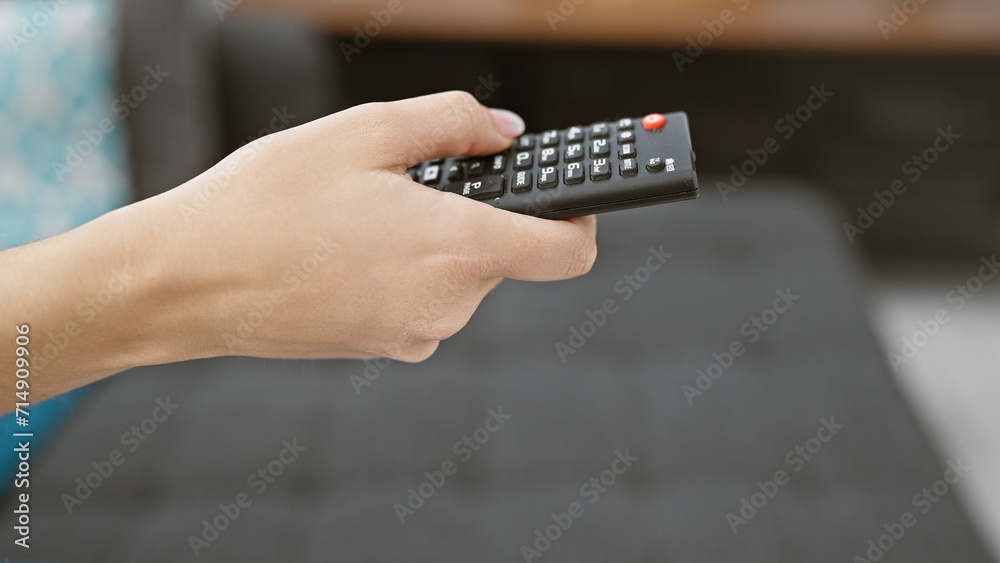 Close-up of a person's hand holding a black television remote control in a blurred indoor living space.