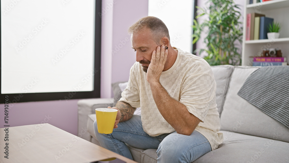 Worried young man, sipping morning coffee at home, deep in thought on his living room sofa.