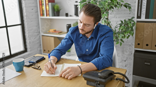 Young hispanic man business worker listening to music taking notes smiling at the office