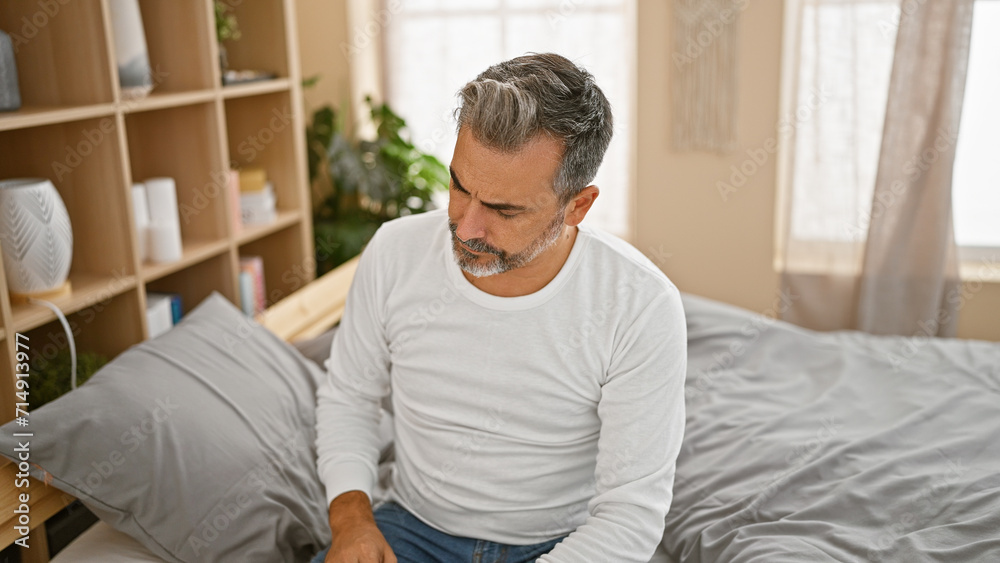Handsome, young hispanic man with grey hair, showing a serious expression while sitting on a comfortable bed and intensely thinking, engulfed in worry, in the relaxed indoors of his apartment bedroom. Handsome, young hispanic man with grey hair, showing a serious expression while sitting on a comfortable bed and intensely thinking, engulfed in worry, in the relaxed indoors of his apartment bedroom.