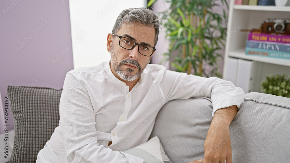 Troubled young hispanic man with grey-haired beard, wearing glasses, reveals a serious expression while sitting on living room sofa, indoors at home. a worrying sign of possible distress. Troubled young hispanic man with grey-haired beard, wearing glasses, reveals a serious expression while sitting on living room sofa, indoors at home. a worrying sign of possible distress.