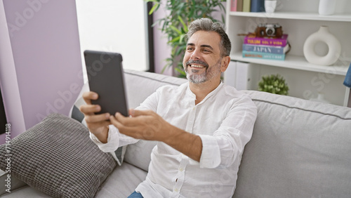 Cheerful young hispanic man with grey hair smiling and having a fun video call on touchpad, relaxing on his living room sofa
