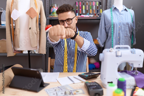 Hispanic man with beard dressmaker designer working at atelier punching fist to fight, aggressive and angry attack, threat and violence