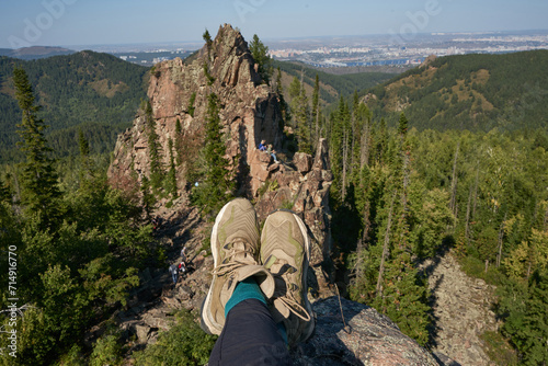 Traveler resting on top of a cliff Igneous rock formation. Summer natural landscape, Stolby national park, Human legs in hiking boots. Hills in the forest. Krasnoyarsk city, Siberia, Russia. Lifestyle