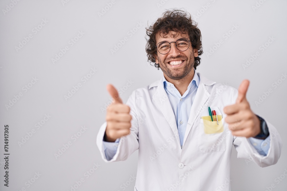 Hispanic young man wearing doctor uniform success sign doing positive gesture with hand, thumbs up smiling and happy. cheerful expression and winner gesture. Hispanic young man wearing doctor uniform success sign doing positive gesture with hand, thumbs up smiling and happy. cheerful expression and winner gesture.