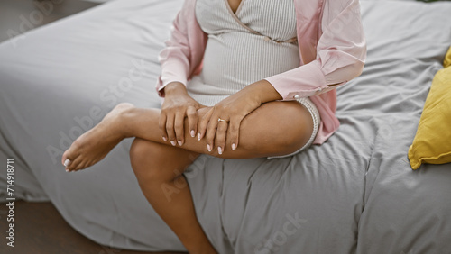 Exhausted young hispanic woman expecting motherhood, sitting on bed, indoors, massaging tired leg in her cosy bedroom, touching pregnant belly in pyjamas.