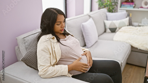 Serious-faced, pregnant young woman sitting at home, casually touching her belly on her living room sofa, embodying the emotion and relaxed anticipation of motherhood.