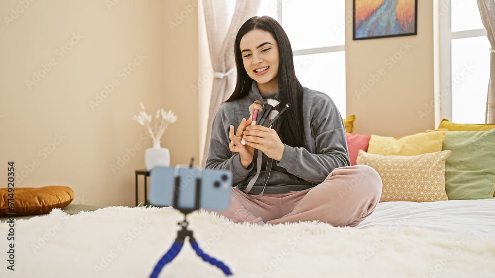 A beautiful hispanic woman records a makeup tutorial in her cozy bedroom with a smartphone on a tripod. A beautiful hispanic woman records a makeup tutorial in her cozy bedroom with a smartphone on a tripod.