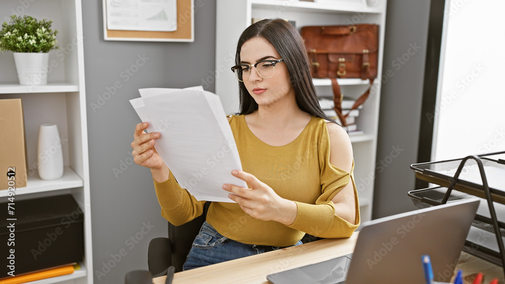 Professional hispanic woman in glasses attentively reading documents at her modern office workspace. Professional hispanic woman in glasses attentively reading documents at her modern office workspace.
