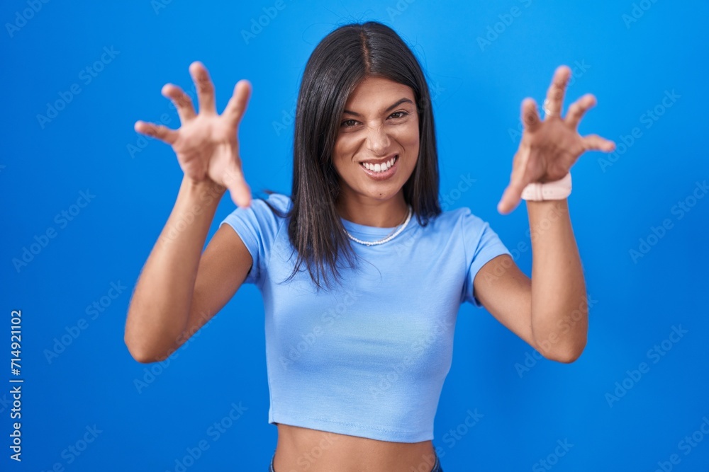 Brunette young woman standing over blue background smiling funny doing claw gesture as cat, aggressive and sexy expression Brunette young woman standing over blue background smiling funny doing claw gesture as cat, aggressive and sexy expression