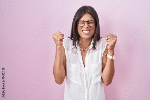 Brunette young woman standing over pink background wearing glasses excited for success with arms raised and eyes closed celebrating victory smiling. winner concept.
