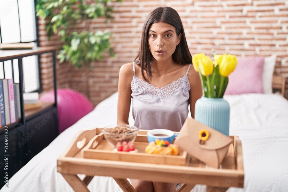 Brunette young woman eating breakfast sitting on the bed scared and amazed with open mouth for surprise, disbelief face