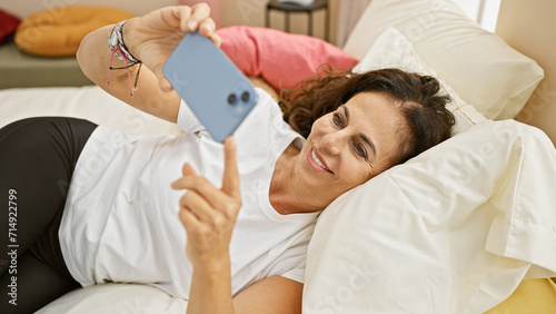 Smiling middle-aged hispanic woman using smartphone in a cozy bedroom setting.