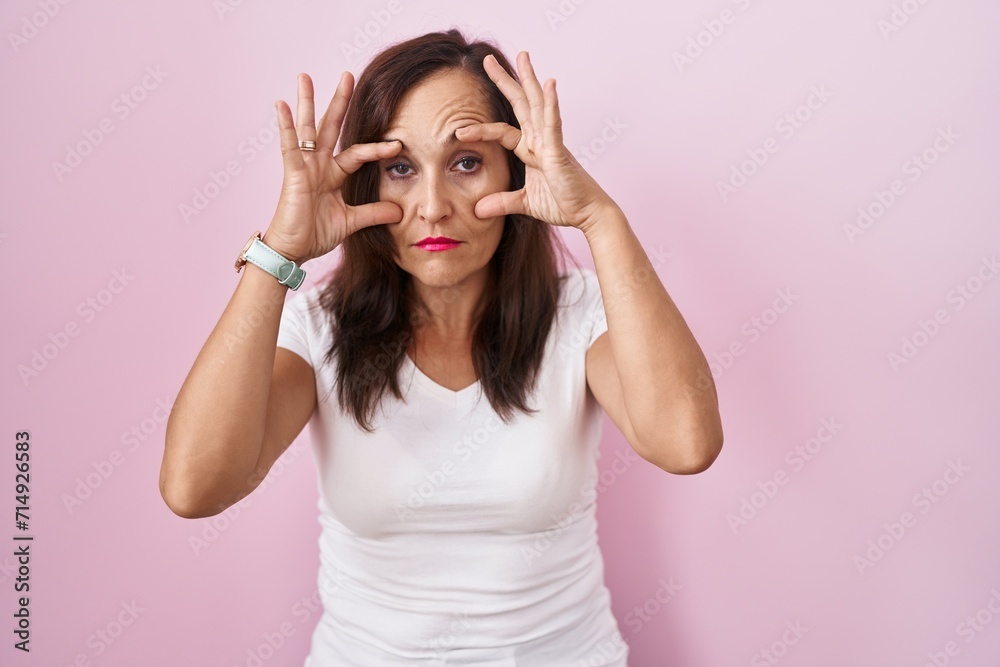 Middle age brunette woman standing over pink background trying to open eyes with fingers, sleepy and tired for morning fatigue