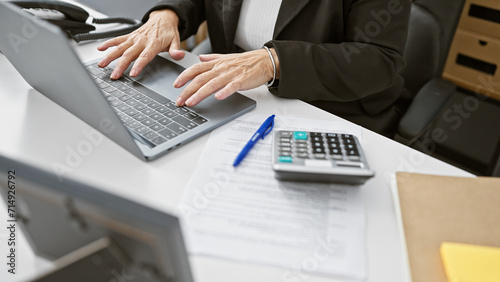 A professional woman working on her laptop in an office with documents, a pen, and calculator present.