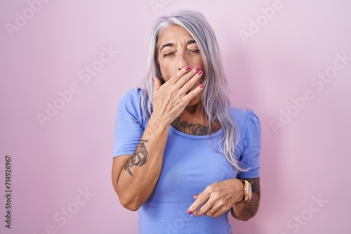 Middle age woman with tattoos standing over pink background bored yawning tired covering mouth with hand. restless and sleepiness.