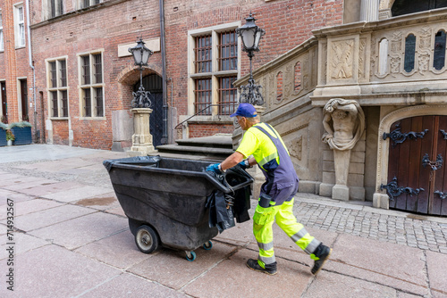 Photography A worker pushes a wastebasket on a city street against a background of building