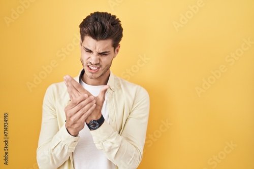 Young hispanic man standing over yellow background suffering pain on hands and fingers, arthritis inflammation